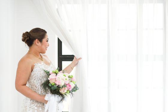 A bride drawing back a sheer curtain in window light